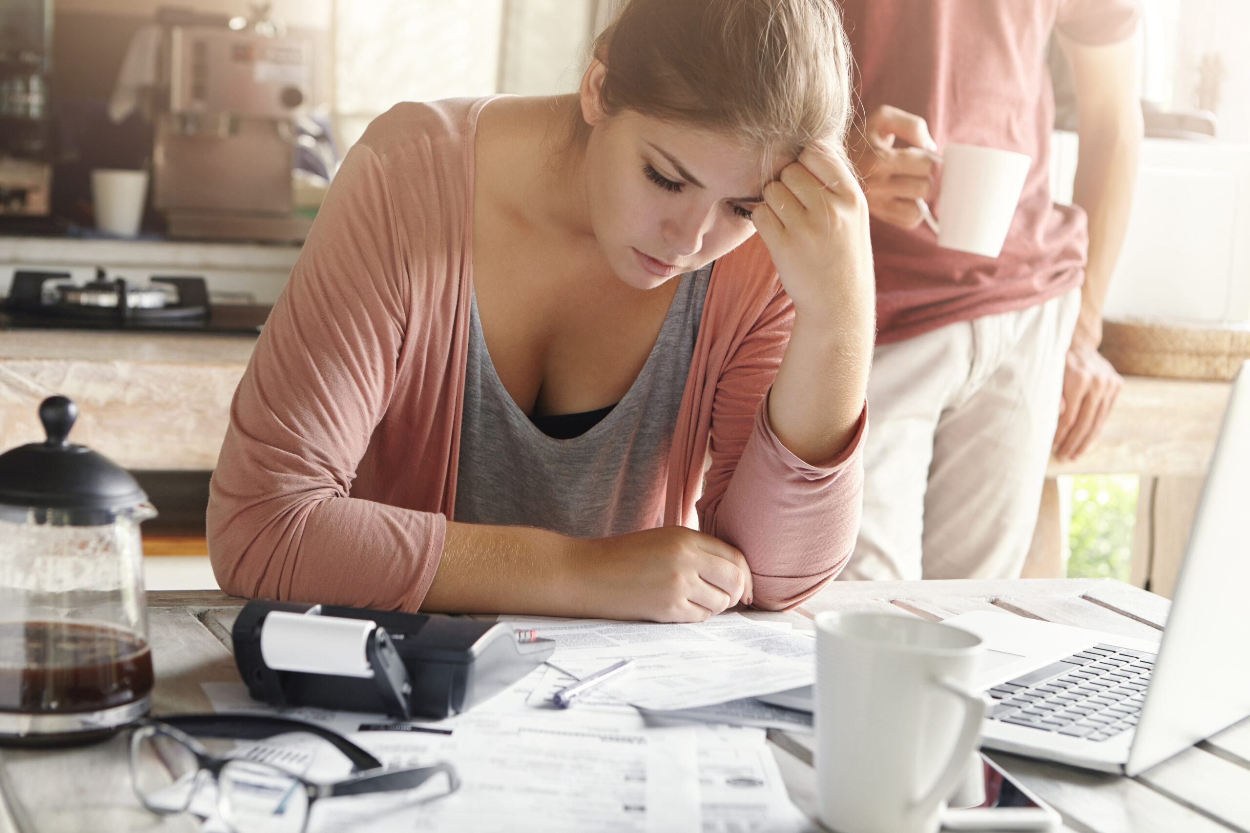Young casual female having depressed look while managing family finances and doing paperwork, sitting at kitchen table with lots of papers, calculator and laptop, her husband standing on background